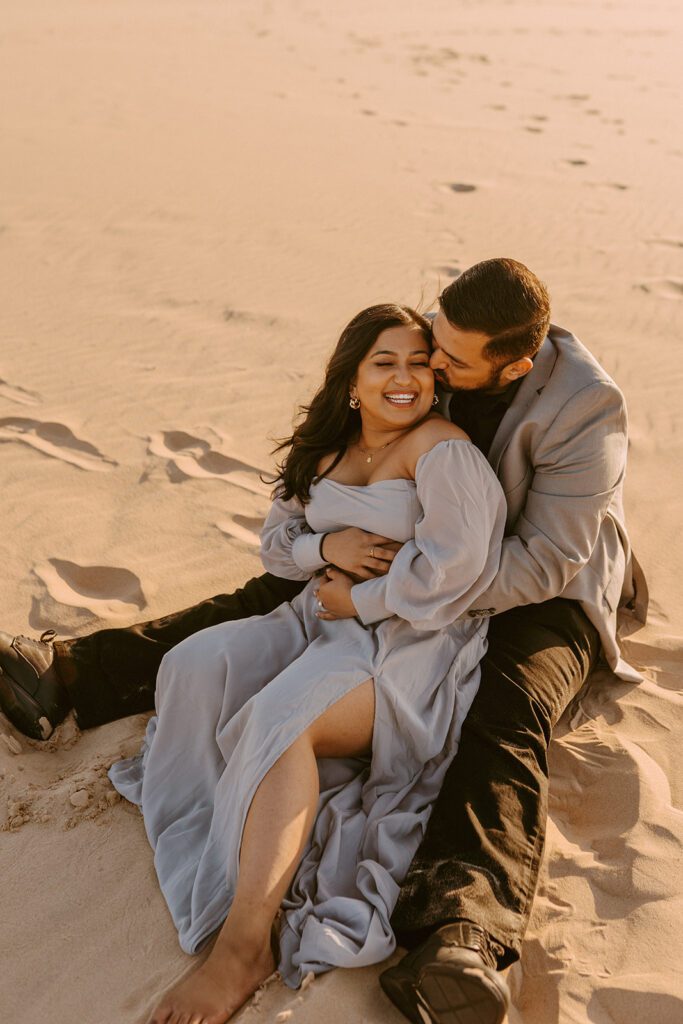 couple sits in sand at their silver lake sand dunes engagement session